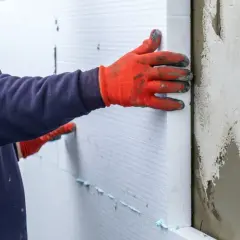 Ręce w pomarańczowych rękawicach przyklejają biały styropian do ściany piwnicy.