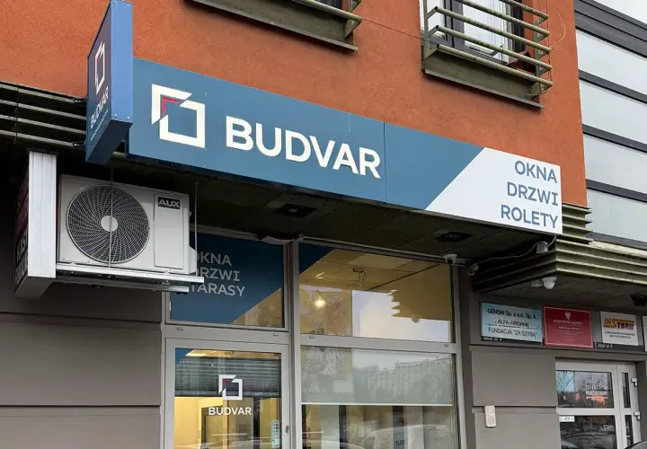 Logo firmy BUDVAR z napisem "OKNA DRZWI ROLETY" na fasadzie budynku.