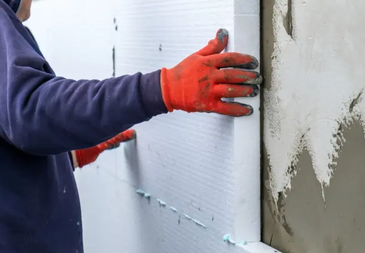 Ręce w pomarańczowych rękawicach przyklejają biały styropian do ściany piwnicy.
