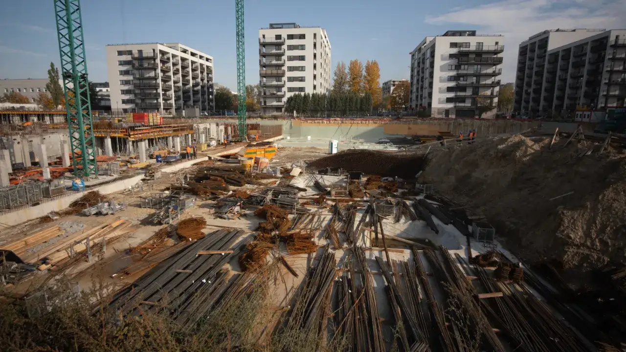 Plac budowy z żurawiem, zbrojeniem i budynkami mieszkalnymi w tle.